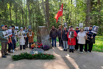 Участники акции «Бессмертный полк» стоят в лесу перед небольшим мемориалом. Многие держат в руках портреты в рамках на палках, а у одного из них развевается красный флаг с серпом и молотом.