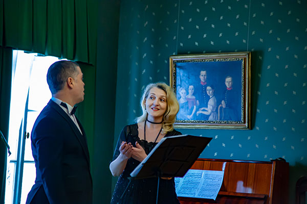 Валерия Дунюшкина играет на черном электронном пианино. Марина Мариненко читает с листа бумаги, который она держит в руках. На стене позади женщин висят две картины.